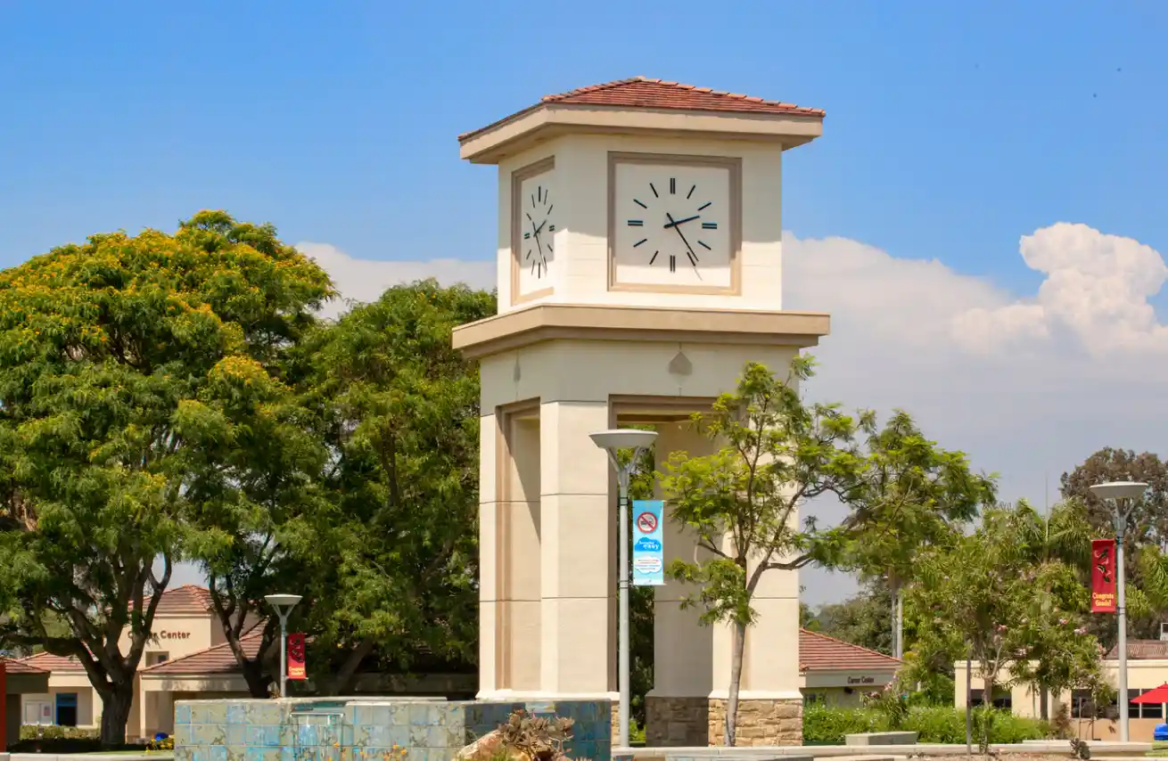Tall clock tower with a tiled roof surrounded by trees, lamp posts, and buildings under a blue sky.