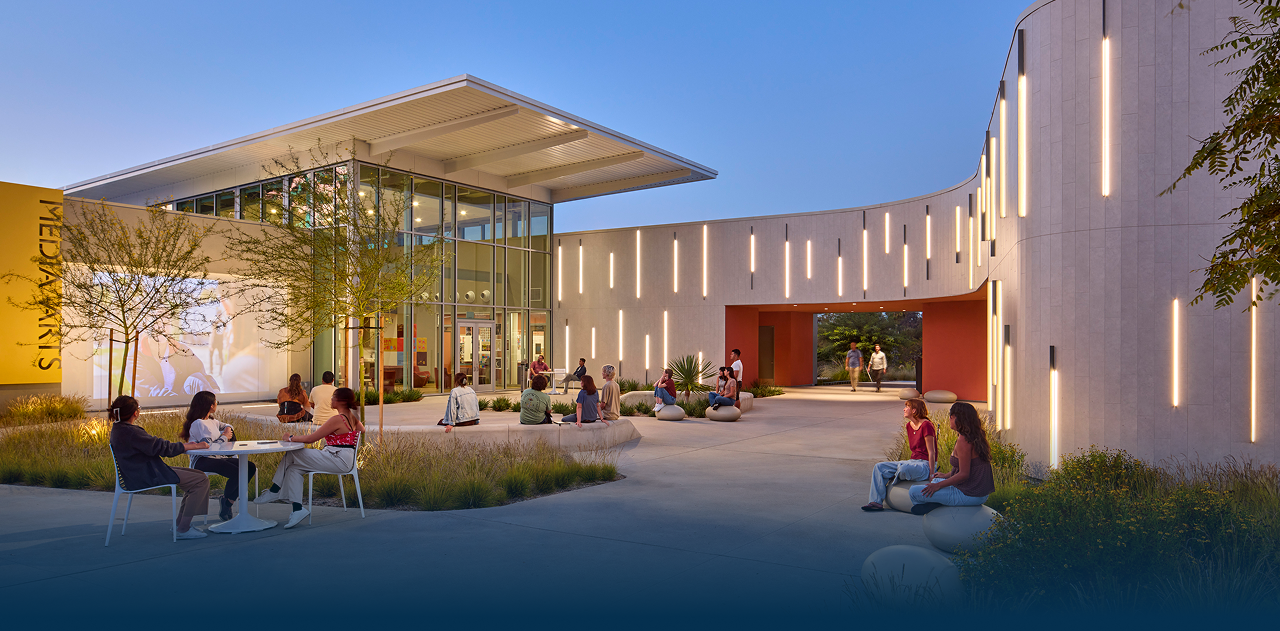 Outdoor courtyard with people seated and walking by a modern building illuminated with vertical lights at dusk.
