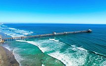 A long pier extending into the ocean over waves near a sandy beach under a clear blue sky.