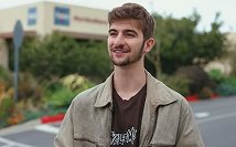 Young man with beard and jacket standing outdoors with blurred greenery and buildings in the background.