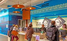 People interact at a colorful reception desk with a vibrant mural and bright ceiling lamps.