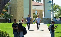 People walking and sitting in a sunny outdoor area with grass, trees, and buildings.